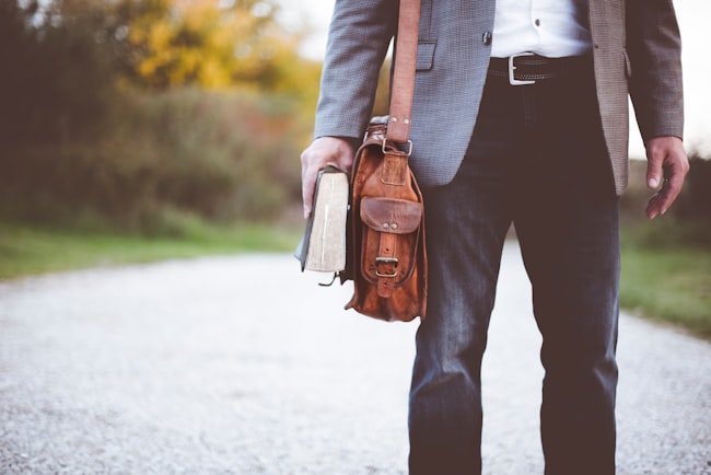 Um homem caminhando em uma estrada vazia vestido de forma social com uma bolsa de couro marrom e um livro