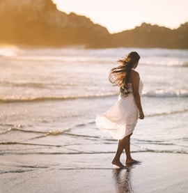 selective focus photo of woman standing on sea shore near rock formation during golden hour