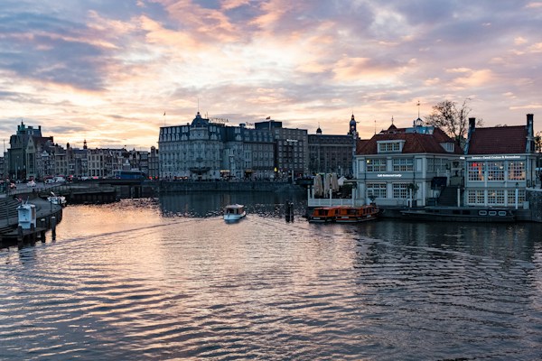 A waterfront scene at sunset with a cityscape featuring historic buildings. A canal is in the foreground, with a small boat moving along the water. The architecture includes prominent, multistory structures with gabled roofs and a larger building displaying a sign that reads 'Victoria'. The sky is filled with vibrant colors from the setting sun, casting reflections on the water.