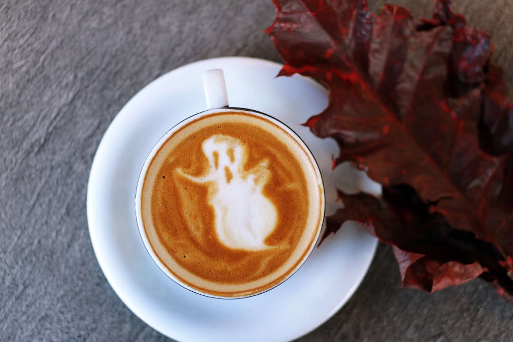 taza de té de cerámica blanca llena de café con leche de ilustración fantasma en platillo de cerámica blanca junto a la fotografía de hoja granate