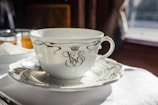 A white porcelain teacup with elegant black decorative patterns and a crown symbol sits on a matching saucer. A metal spoon rests on the saucer. In the background, there are out-of-focus jars with lids, likely containing spreads or condiments. Soft lighting suggests a cozy and refined setting.