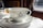 A white porcelain teacup with elegant black decorative patterns and a crown symbol sits on a matching saucer. A metal spoon rests on the saucer. In the background, there are out-of-focus jars with lids, likely containing spreads or condiments. Soft lighting suggests a cozy and refined setting.