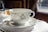 A white porcelain teacup with elegant black decorative patterns and a crown symbol sits on a matching saucer. A metal spoon rests on the saucer. In the background, there are out-of-focus jars with lids, likely containing spreads or condiments. Soft lighting suggests a cozy and refined setting.