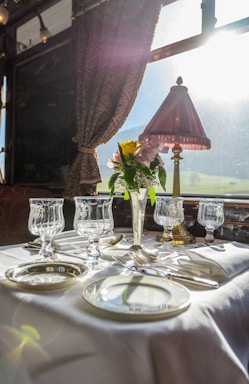 A luxurious dining setting on a train features a beautifully set table with elegant glassware and silverware. A vase with fresh flowers adds a touch of color, and a classic table lamp casts a warm glow. The sunlight streams through the large window, enhancing the bright and sophisticated atmosphere.