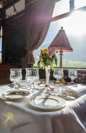 A luxurious dining setting on a train features a beautifully set table with elegant glassware and silverware. A vase with fresh flowers adds a touch of color, and a classic table lamp casts a warm glow. The sunlight streams through the large window, enhancing the bright and sophisticated atmosphere.