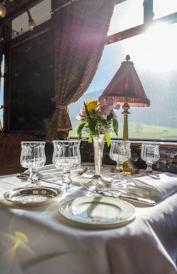 A luxurious dining setting on a train features a beautifully set table with elegant glassware and silverware. A vase with fresh flowers adds a touch of color, and a classic table lamp casts a warm glow. The sunlight streams through the large window, enhancing the bright and sophisticated atmosphere.