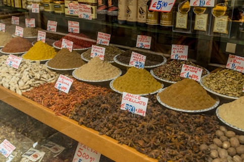 A market display featuring a variety of spices arranged in piles on plates, each labeled with handwritten tags and prices. The spices include vibrant colors such as yellow, red, and brown.