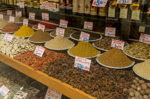 A market display featuring a variety of spices arranged in piles on plates, each labeled with handwritten tags and prices. The spices include vibrant colors such as yellow, red, and brown.