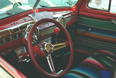 The interior of a vintage car is depicted, featuring a red steering wheel and chromed dashboard with classic controls and knobs. The seats and door panels are upholstered with colorful striped fabric.