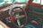 The interior of a vintage car is depicted, featuring a red steering wheel and chromed dashboard with classic controls and knobs. The seats and door panels are upholstered with colorful striped fabric.