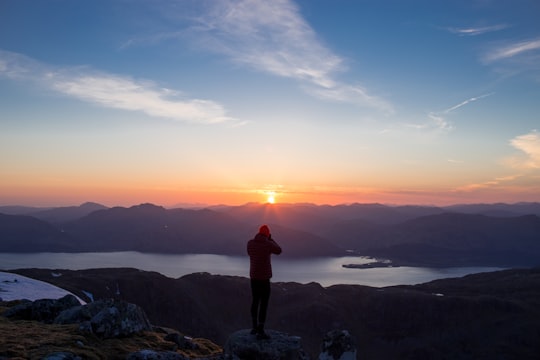 person standing on mountain ridge over lake under cloudy sky at sunset in Ballachulish United Kingdom