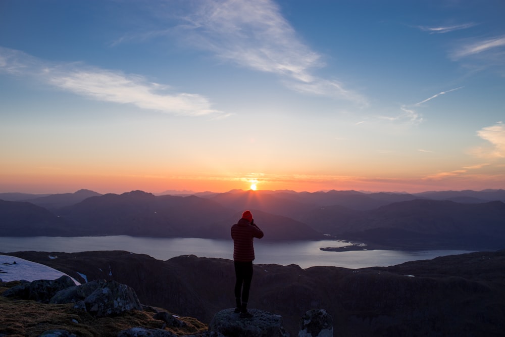 person standing on mountain ridge over lake under cloudy sky at sunset