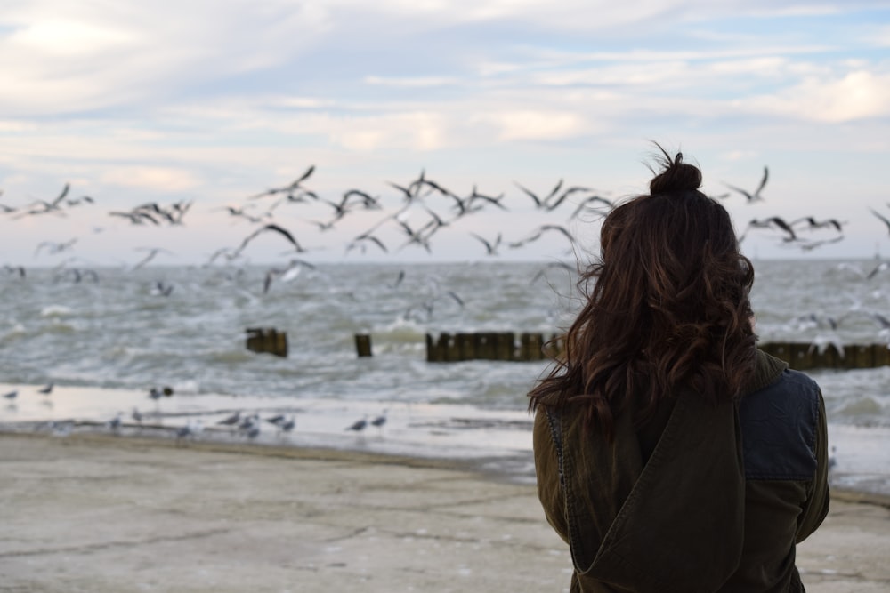 woman looking at flock of birds flying over body of water under cloudy sky