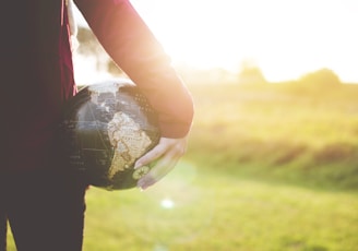 person holding black and brown globe ball while standing on grass land golden hour photography