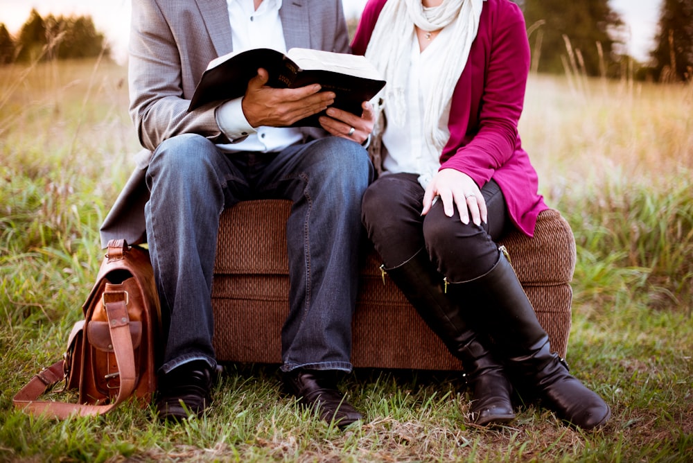 A married couple sitting on an ottoman outdoors. He is reading a book.