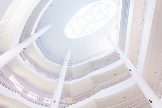 A luminous, spacious interior space with a circular architectural design featuring multiple ascending levels and a large skylight. The design is modern, with white walls and sleek lines, creating an airy and open atmosphere.