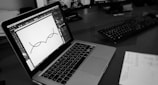 A black and white image of a laptop on a desk displaying a design software with a simple line illustration. There are two monitors in the background and a keyboard on the desk. Papers with printed text are partially visible in the foreground.