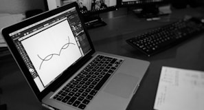 A black and white image of a laptop on a desk displaying a design software with a simple line illustration. There are two monitors in the background and a keyboard on the desk. Papers with printed text are partially visible in the foreground.