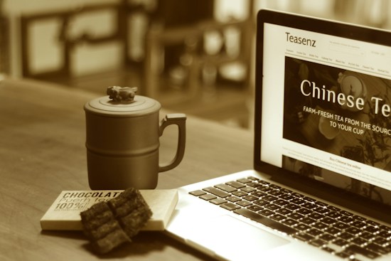 A rustic mug with a lid sits on a wooden surface next to a partially open laptop displaying a website about Chinese tea. In front of the mug, there is a chocolate bar with some pieces broken off and placed nearby. The scene is warm and cozy, lit in sepia tones.