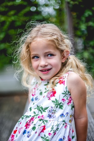 A young girl with curly blonde hair wearing a white dress adorned with colorful floral patterns. She has an amused and slightly mischievous expression, set against a blurred, leafy background.