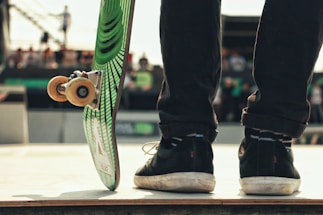 A skateboarder stands on a ramp, with the focus on their shoes and skateboard. The skateboard is propped up, showing its underside design with green and black stripes. The background is slightly blurred, suggesting an outdoor event with spectators.