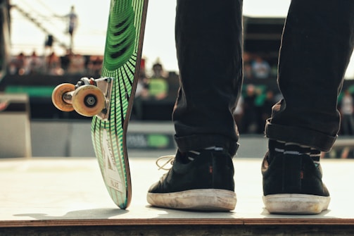 A skateboarder stands on a ramp, with the focus on their shoes and skateboard. The skateboard is propped up, showing its underside design with green and black stripes. The background is slightly blurred, suggesting an outdoor event with spectators.
