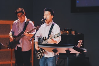 Two musicians are performing on stage, each playing a guitar. The setting has a cozy, indoor atmosphere with soft lighting. One musician is singing into a microphone while playing an electric guitar, and the other focuses on a bass guitar.