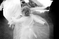 A young child in a flowing, white dress with lace details is seen from behind, holding out their arms in a carefree manner. The scene appears to be set outdoors on a path with soft focus on the background, giving a dreamy, ethereal atmosphere. The ground is unpaved, suggesting a natural setting.