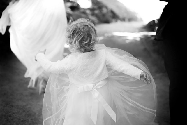 A young child in a flowing, white dress with lace details is seen from behind, holding out their arms in a carefree manner. The scene appears to be set outdoors on a path with soft focus on the background, giving a dreamy, ethereal atmosphere. The ground is unpaved, suggesting a natural setting.
