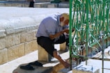 An elderly man with a white beard leans forward to wash his hands at an outdoor faucet. He wears a striped shirt and black pants, and his cap is knitted. His sandals are placed beside him. The setting is a stone courtyard with a decorative green metal fence nearby.