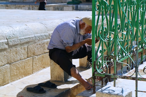 An elderly man with a white beard leans forward to wash his hands at an outdoor faucet. He wears a striped shirt and black pants, and his cap is knitted. His sandals are placed beside him. The setting is a stone courtyard with a decorative green metal fence nearby.