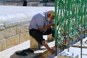 An elderly man with a white beard leans forward to wash his hands at an outdoor faucet. He wears a striped shirt and black pants, and his cap is knitted. His sandals are placed beside him. The setting is a stone courtyard with a decorative green metal fence nearby.