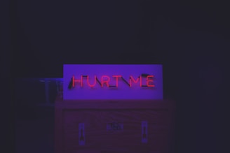 Red neon "Hurt Me" sign on top of a crate with arrow stickers