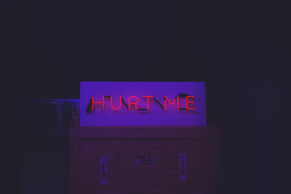 Red neon "Hurt Me" sign on top of a crate with arrow stickers