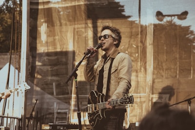 A musician wearing sunglasses is performing on stage, holding an electric guitar and singing into a microphone. The stage is set outdoors with a backdrop that has a semi-transparent, patterned curtain. The musician is dressed in a light-colored jacket and dark pants. The atmosphere appears casual and relaxed, typical of an outdoor concert.