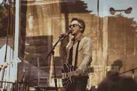 A musician wearing sunglasses is performing on stage, holding an electric guitar and singing into a microphone. The stage is set outdoors with a backdrop that has a semi-transparent, patterned curtain. The musician is dressed in a light-colored jacket and dark pants. The atmosphere appears casual and relaxed, typical of an outdoor concert.