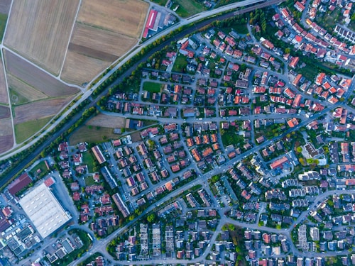 Aerial view of a suburban area with a grid of streets and small houses. There are open fields on one side and a mix of residential buildings and some larger warehouses or industrial buildings. The area is intersected by what appears to be railway tracks or a highway.