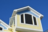 The top portion of a house with light yellow siding and a blue, fish scale-patterned accent under the gable roof is visible. The roof is white-trimmed and features an arched window, with an open pane slightly ajar, all set against a clear blue sky.
