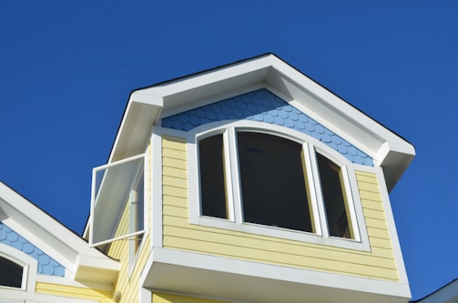 The top portion of a house with light yellow siding and a blue, fish scale-patterned accent under the gable roof is visible. The roof is white-trimmed and features an arched window, with an open pane slightly ajar, all set against a clear blue sky.
