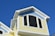 The top portion of a house with light yellow siding and a blue, fish scale-patterned accent under the gable roof is visible. The roof is white-trimmed and features an arched window, with an open pane slightly ajar, all set against a clear blue sky.