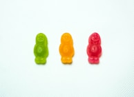 Three gummy candies in the shapes of smiling figures are arranged horizontally, each in a different color: green, orange, and red, set against a textured white background.