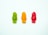 Three gummy candies in the shapes of smiling figures are arranged horizontally, each in a different color: green, orange, and red, set against a textured white background.