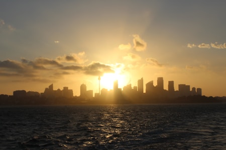 A skyline is silhouetted against a vibrant sunset, with the sun partially hidden behind the buildings. The sky is filled with warm tones of orange and yellow, with clouds adding depth to the scene. The water in the foreground reflects the colors of the sky, creating a serene and picturesque view.
