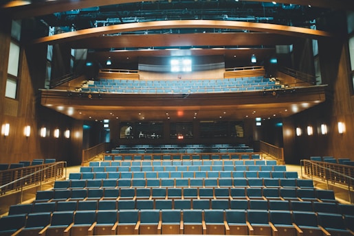 A spacious, empty auditorium with neatly arranged rows of blue seats and wooden accents throughout. The room features a balcony section above, lined with additional seating. Soft lighting creates a warm atmosphere, and the ceiling includes exposed beams and lighting fixtures.