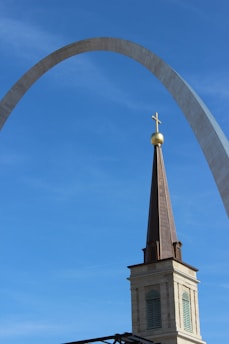 A tall church steeple topped with a cross is set against a blue sky. Behind it, the iconic Gateway Arch is visible, creating an interesting juxtaposition between the two structures.