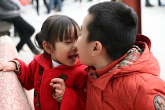 Two children are playfully interacting, with one child wearing a red coat and the other in a red jacket with a plaid inner lining. They are at a public place, surrounded by other people, with a patterned railing on the side.