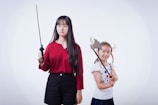 Two girls are posing with props. The older girl, wearing a red top and black shorts, is holding a sword with a serious expression. The younger girl, wearing a polka-dotted shirt and a Viking helmet with pigtail braids, is holding an axe prop while smiling.