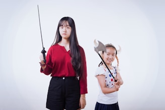 Two girls are posing with props. The older girl, wearing a red top and black shorts, is holding a sword with a serious expression. The younger girl, wearing a polka-dotted shirt and a Viking helmet with pigtail braids, is holding an axe prop while smiling.