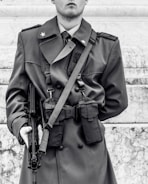 A person is wearing a military-style coat with shoulder epaulets and star insignia. They are holding a firearm in their right hand. The coat has pockets and appears to be well-fitted. The background consists of marble steps.