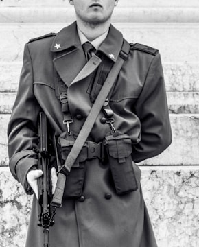A person is wearing a military-style coat with shoulder epaulets and star insignia. They are holding a firearm in their right hand. The coat has pockets and appears to be well-fitted. The background consists of marble steps.